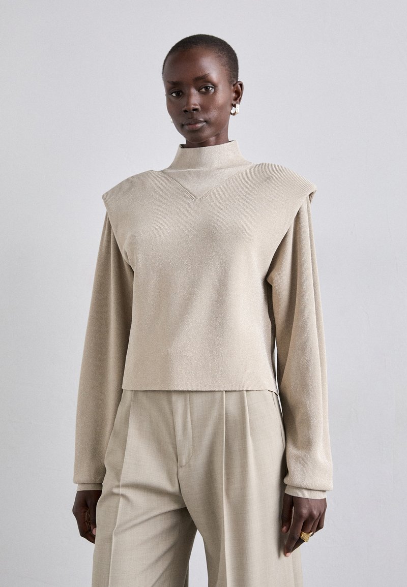 Beige metallisk strikket pullover med høj hals og strukturerede skuldre, parret med lyse beige plisserede bukser. Glat tekstur, afslappet pasform.
