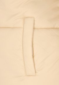 Detaliu al unei geacă puffer bej cu un finisaj moale, texturat și un design cu buzunare verticale. Coasăt pe margini pentru o structură suplimentară.