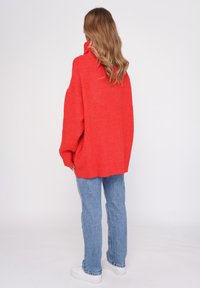 Przesadnie duży czerwony sweter robiony na drutach z wysokim kołnierzem, teksturowana tkanina i opuszczone ramiona, w połączeniu z jasnoniebieskimi spodniami typu straight-leg oraz białymi butami.