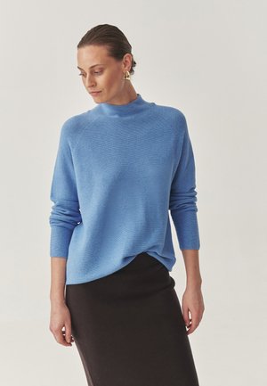 Lyseblå strikket sweater med høj hals og lange ærmer, parret med en tætsiddende mørkebrun nederdel, der har en glat tekstur og en afslappet pasform.