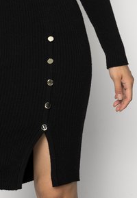 Černé žebrované šaty s dlouhými rukávy a bočním rozparkem, s pěti kovovými knoflíky podél švu pro dodatečný detail.