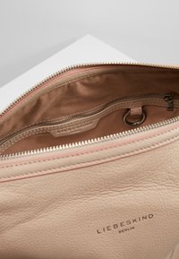 Wnętrze torby ze jasnobeżowej skóry o gładkiej teksturze, z kieszenią na zamek i metką z logo. Posiada kontrastujące szczegóły w różowym kolorze, w tym zamek błyskawiczny i przeszycia.