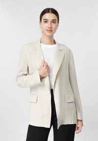 Femme aux cheveux bruns attachés en chignon bas, portant un blazer beige sur une chemise blanche et un pantalon noir, debout devant un fond clair uni.