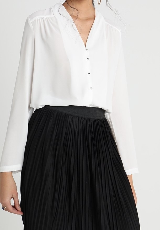 Femme portant un chemisier blanc à manches longues avec des boutons et une jupe plissée noire taille haute.