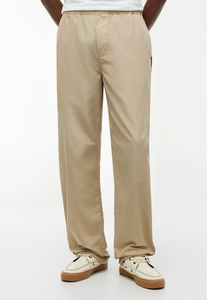 Person, die beige locker sitzende Hosen und weiße Schnür-Sneaker mit braunen Sohlen trägt, vor einem schlichten weißen Hintergrund stehend.