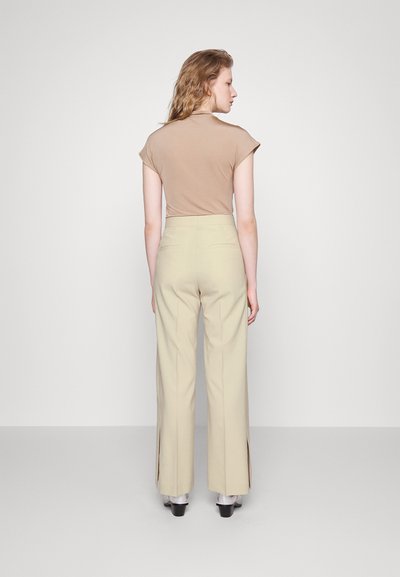 Woman standing facing away, wearing beige high-waisted pants with side slits and a light brown short-sleeve top on plain background.