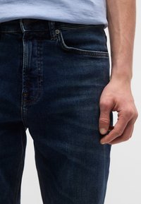 Σκούρο μπλε τζιν από denim, με μπροστινή τσέπη, ραφές σε αντίθεση και φερμουάρ με μεταλλικό κουμπί.