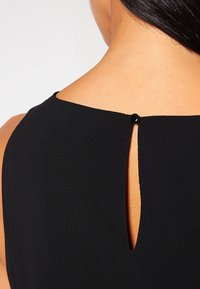 Close-up van een zwart mouwloos kledingstuk dat met een kleine zwarte knoop aan de achterkant van de halslijn op blote huid is vastgemaakt.