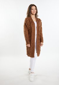 Brun strikket cardigan med kabelmønster, åben foran design, lange ærmer, parret med hvide bukser og sneakers på en neutral baggrund.
