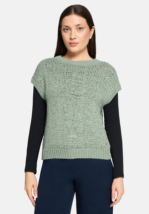 Mintgroene gebreide trui met korte mouwen, gestructureerd ontwerp, geribbelde zoom, gedragen over een zwart langarmshirt, gecombineerd met donkere broek.