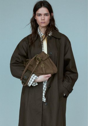 Jong persoon met lang donker haar draagt een oversized bruine jas, geruit overhemd en houdt een bijpassende bruine leren handtas vast tegen een effen achtergrond.