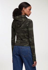 Felpa a maniche lunghe verde mimetica con design aderente, tessuto texturizzato e cappuccio. Abbinata a jeans blu a vita alta.