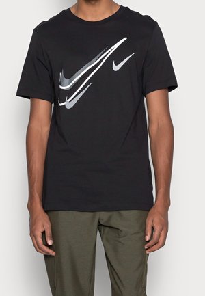 Mężczyzna w czarnej koszulce z wieloma nakładającymi się białymi i szarymi logo Nike w kształcie łyżwy, w połączeniu z oliwkowozielonymi spodniami.
