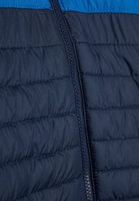 Marineblauwe doorgestikte pufferjack met een soepele rits, horizontale stiksels en een contrasterend blauwe bovenpaneel. Zachte textuur.