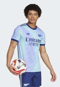 Lyseblå fodboldtrøje med gradientdesign, marineblå krave og detaljer. Har teksten "Emirates Fly Better" og Adidas-logo. Spiller holder en bold.