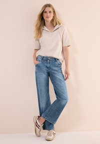 Sweatshirt à capuche beige clair avec des manches courtes, associé à un pantalon en denim bleu à jambes larges. L'ensemble est complété par des baskets claires avec des accents bordeaux.