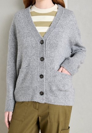 Femme portant un cardigan gris boutonné avec des poches avant sur une chemise beige rayée et un pantalon vert olive, main dans la poche du cardigan.