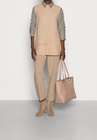Gilet en tricot sans manches beige sur une chemise à carreaux, associé à un pantalon beige et des bottines taupe, tenant un sac fourre-tout rose pâle.
