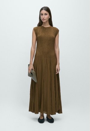 Kobieta ubrana w brązową plisowaną sukienkę maxi bez rękawów, trzymająca szarą, teksturowaną kopertówkę, ubrana w czarne baleriny, stojąca na jednolitym tle.