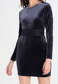 Sjöblå sammetsklänning med figursydd silhuett, långa ärmar med spetskant och en bred spetssnar för detaljer i midjan. Mjuk textur och slät finish.