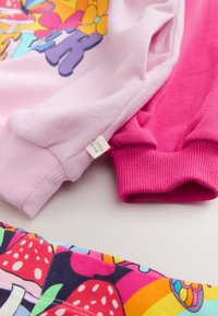 Két pulóver rózsaszín és világos rózsaszín pamutból, bordázott mandzsettákkal, az előtérben virágokat és gombákat ábrázoló színes mintás szövettel.