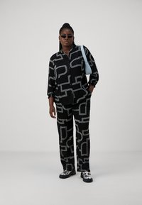 Conjunto negro con un diseño estampado y patrones geométricos blancos. Camisa de botón de manga larga y pantalones a juego. Zapatos plateados, bolso minimalista y gafas de sol.