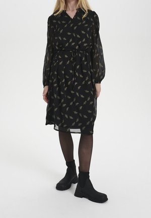 Woman wearing a black knee-length dress with leaf patterns, sheer long sleeves, black tights, and black ankle boots, standing on a white background.