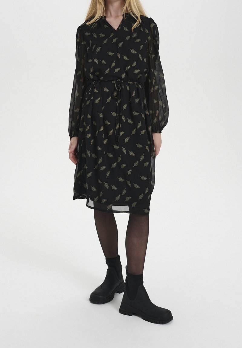 Femme portant une robe noire longueur genoux avec des motifs de feuilles, manches longues transparentes, collants noirs et bottines noires, debout sur un fond blanc.