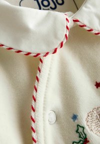 Vêtement de couleur crème avec un tissu doux et texturé. Comprend un col rond avec un bord tordu rouge et blanc et des motifs de fête brodés.