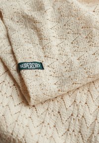 Cremefarvet strikket tekstil med et zigzag-mønster og en struktureret overflade. Har et grønt Superdry mærke i det nederste hjørne.