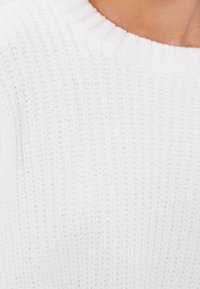 Närbild av en vit stickad tröja med ribbad rund halsringning på en persons övre bröst och nacke.