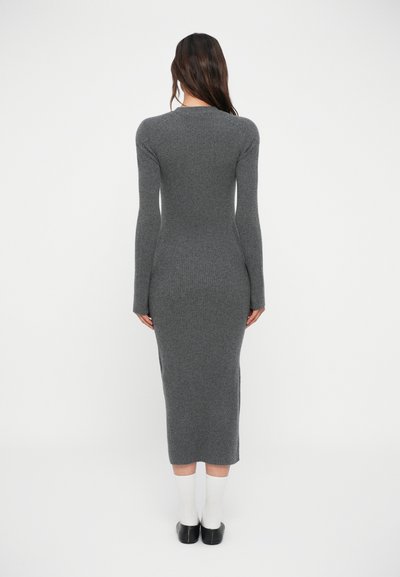 Robe longue grise à manches longues avec une silhouette ajustée, longueur mi-mollet et fentes sur les côtés. Associée à des chaussettes blanches et des chaussures noires.