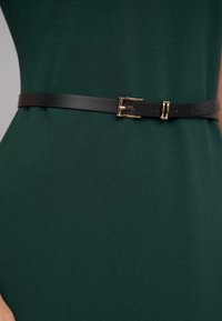Schwarzer Leder-Gürtel mit einer goldfarbenen Schnalle, glatte Textur, entworfen für eine taillierte Passform, getragen über einem dunkelgrünen Kleid aus Stoff.