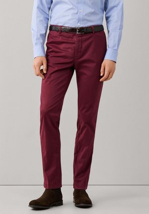 Hombre con pantalones burdeos, cinturón de cuero marrón, camisa de manga larga azul claro a rayas y botas de ante marrones, de pie frente a un fondo liso.