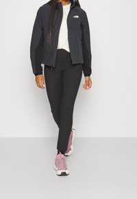 Sort softshelljakke med lynlåslukning, lange ærmer, logo på brystet; kombineret med sorte leggings og lyserøde sneakers.