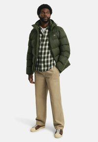Pufferjack in donkergroen, met een gewatteerd ontwerp en een capuchon. Daaronder een geruite shirt in groen en wit. Beige broek en bruine sneakers maken de look compleet.