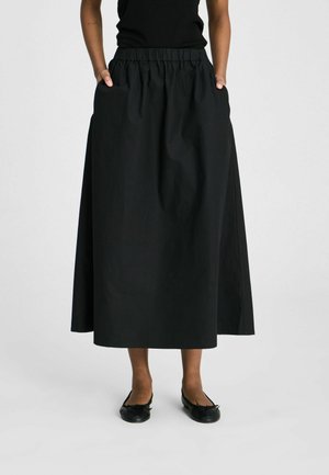 Person trägt einen langen schwarzen Rock mit elastischem Bund und schwarze Ballettschuhe, steht mit den Händen in den Taschen.