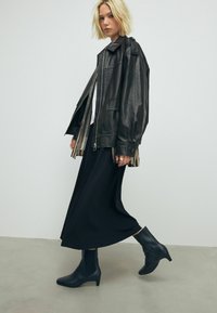 Schwarze Lederübergröße Jacke, weißes Hemd, langer schwarzer Rock, knöchellange marineblaue Stiefel mit spitzer Zehe und einem leichten Absatz, minimalistischem Design.