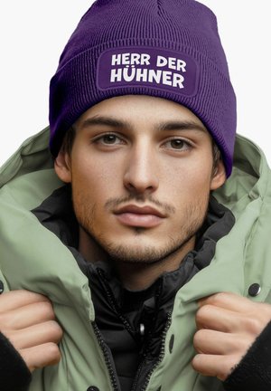 Purple knitted beanie with a white patch featuring text, worn with a green puffer jacket. The jacket has a black interior and high collar.