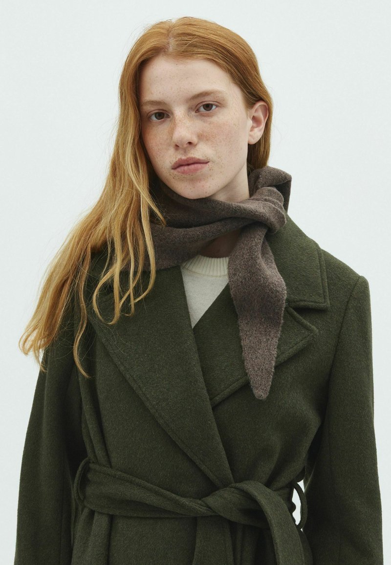 Jeune femme aux longs cheveux roux portant un manteau vert foncé ceinturé et une écharpe marron sur un fond clair uni.