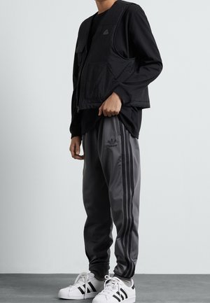 Osoba ubrana w czarną koszulkę z długim rękawem, czarną kamizelkę, szare spodnie dresowe Adidas z czarnymi paskami oraz białe buty sportowe Adidas z czarnymi paskami.