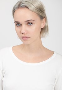 Maglione bianco a maglia con scollatura rotonda, indossato da una persona con capelli corti e chiari e trucco neutro, su uno sfondo semplice.