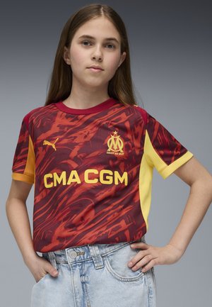 Maglietta rossa e gialla a maniche corte con motivo astratto, caratterizzata da un collo a giro e loghi, abbinata a pantaloni corti di jeans azzurri chiari.