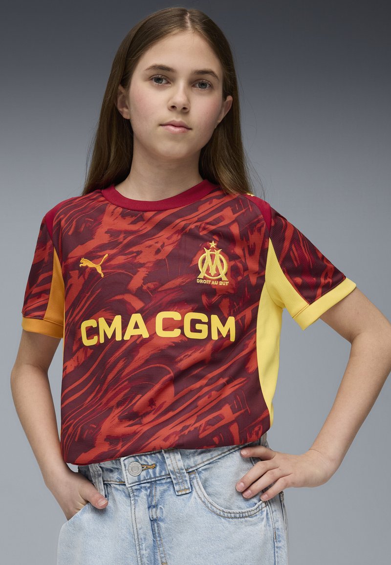 Röd och gul kortärmad tröja med abstrakt mönster, med rundad halsringning och logotyper, tillsammans med ljusblå denimshorts.