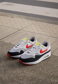 Adidași Nike Air Max în gri, negru și roșu. Caracteristici includ o unitate de aer vizibilă, suede texturat și accente de neon galben pe limbă.