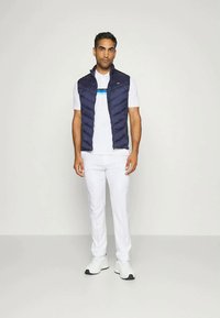 Zils polsterēts bezdelīgas jaka virs baltas t-kreklā ar horizontālu zilu svītru, apvienots ar baltām biksēm un baltām sporta apavām.