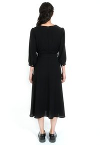 Kobieta z ciemnymi włosami, w niskim półupięciu, ubrana w czarną, midi sukienkę z długim rękawem i guzikami przy mankietach oraz czarne, masywne buty, stojąca plecami.