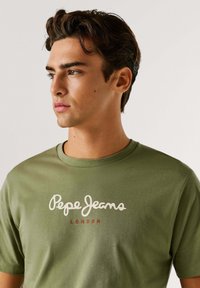 Alyvuogių žalios medvilnės marškinėliai su apvaliu iškirpimu ir spausdintu "Pepe Jeans London" logotipu baltos ir raudonos spalvų ant priekio.
