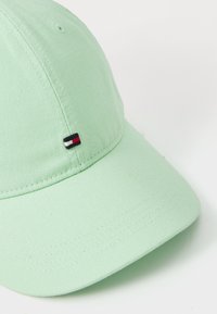 Lys grøn bomulds hat med en buet skygge, der har et lille broderet logo i rød, hvid og sort på den venstre frontside.