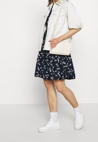 Kvinna i en marinblå blommig klänning, vit jacka, vita sneakers och håller en liten vit vävd crossbody-väska.